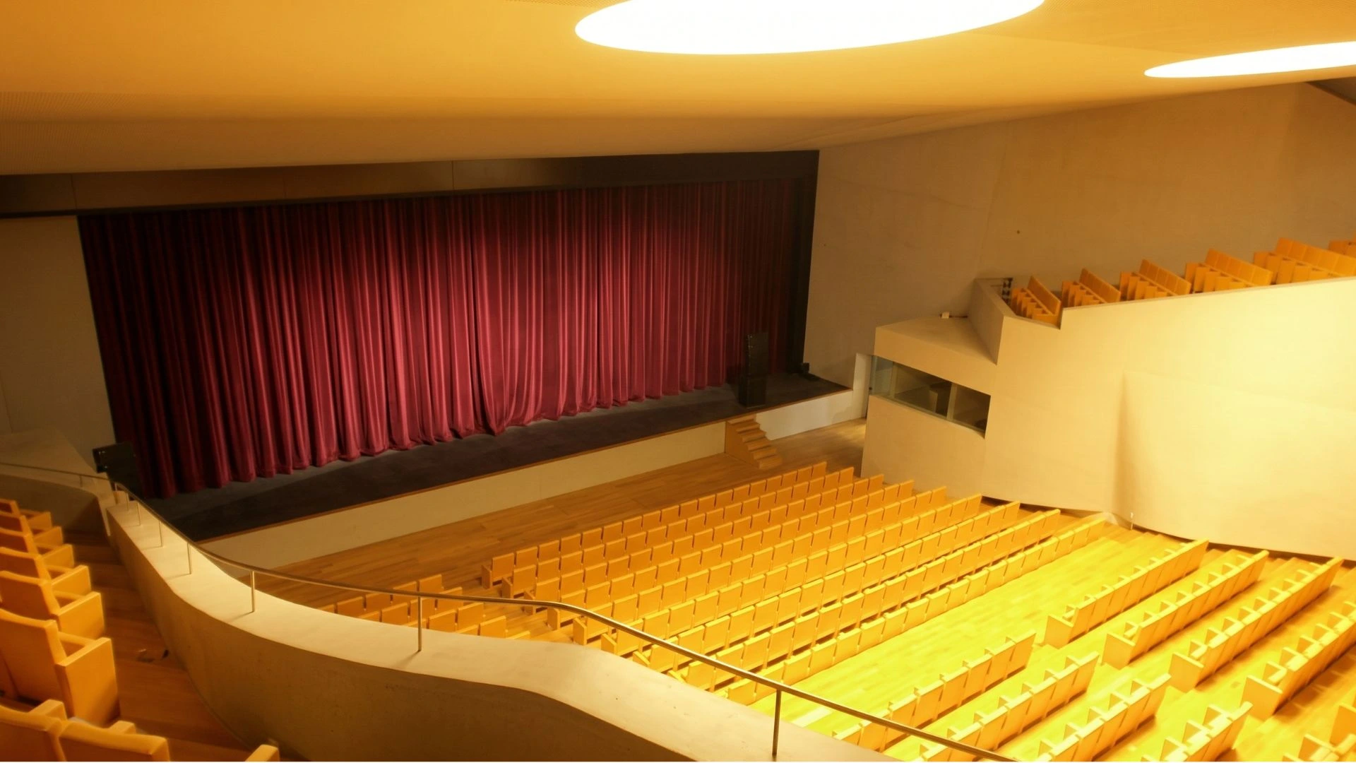 Interior del teatro del Revellín, un teatro moderno e iluminado, con filas de butacas amarillas vacías y el escenario principal cerrado por un gran telón rojo clásico